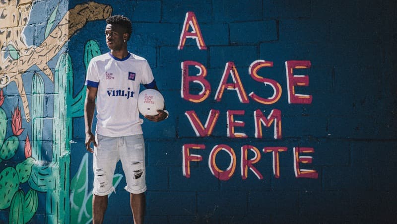 Vini Jr ao lado de muro de seu Instituto escrito "a base vem forte"; Ex-Flamengo tem programa que auxilia na educação de jovens e dá aulas antirracistas