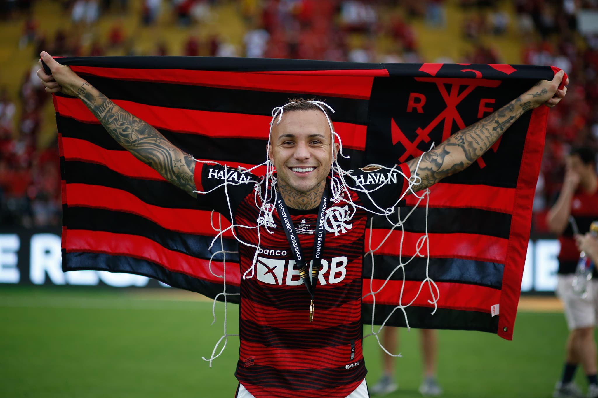Everton Cebolinha segurando bandeira do Flamengo, com pedaço da rede e medalha no pescoço após conquistar libertadores em 2022.