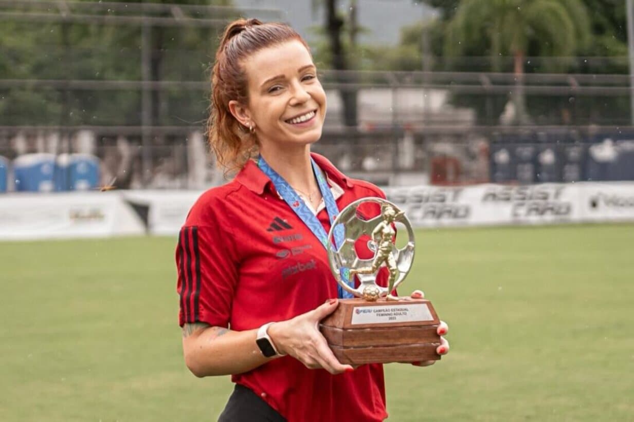 A médica do Flamengo está segurando a taça de campeã carioca e sorrindo, ela está em campo e na chuva