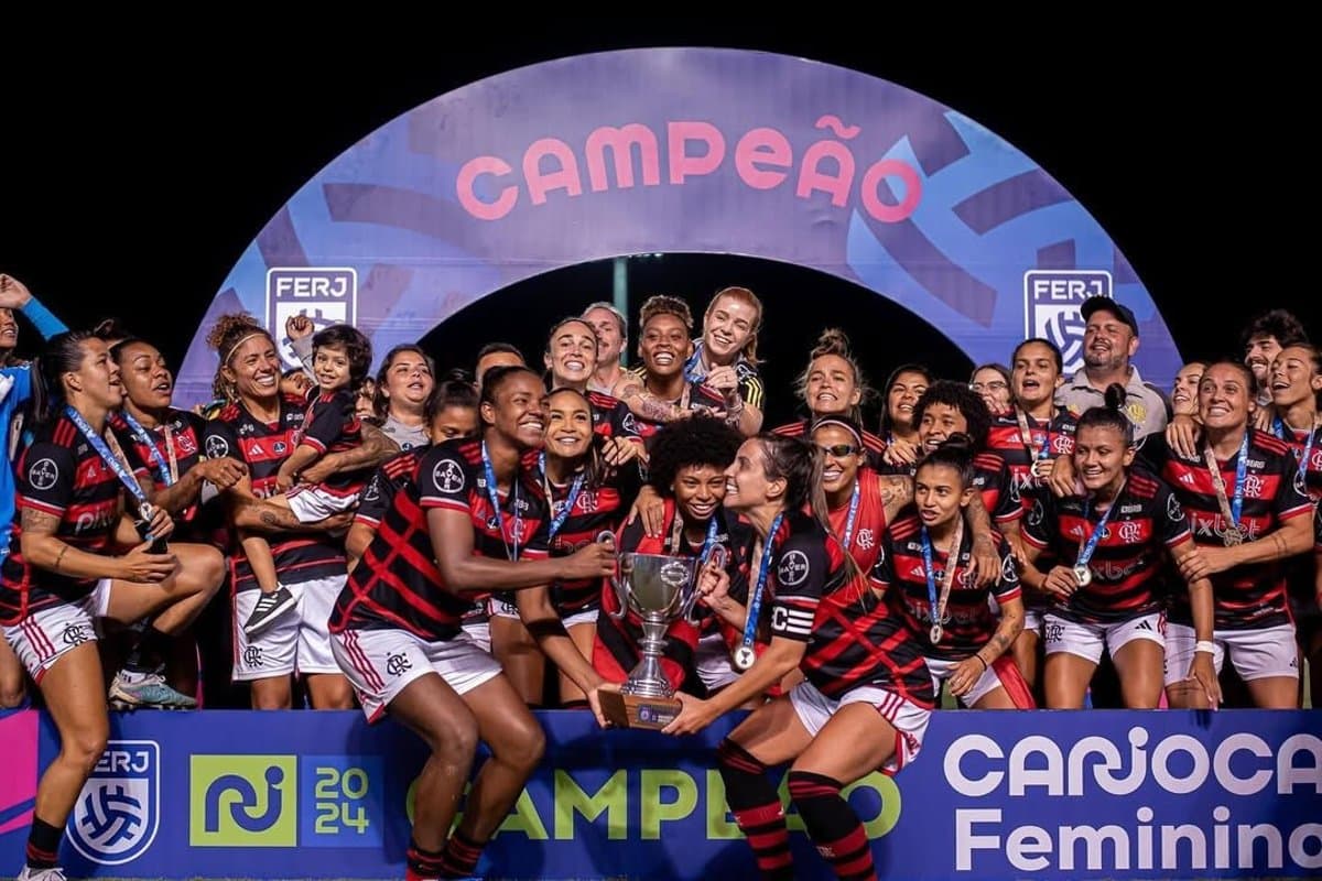 Na imagem o time feminino do Flamengo levanta a Taça do Carioca feminino adulto 2024, as jogadoras estão sorrindo e posicionada em frente ao totem escrito: Campeão Carioca Feminino