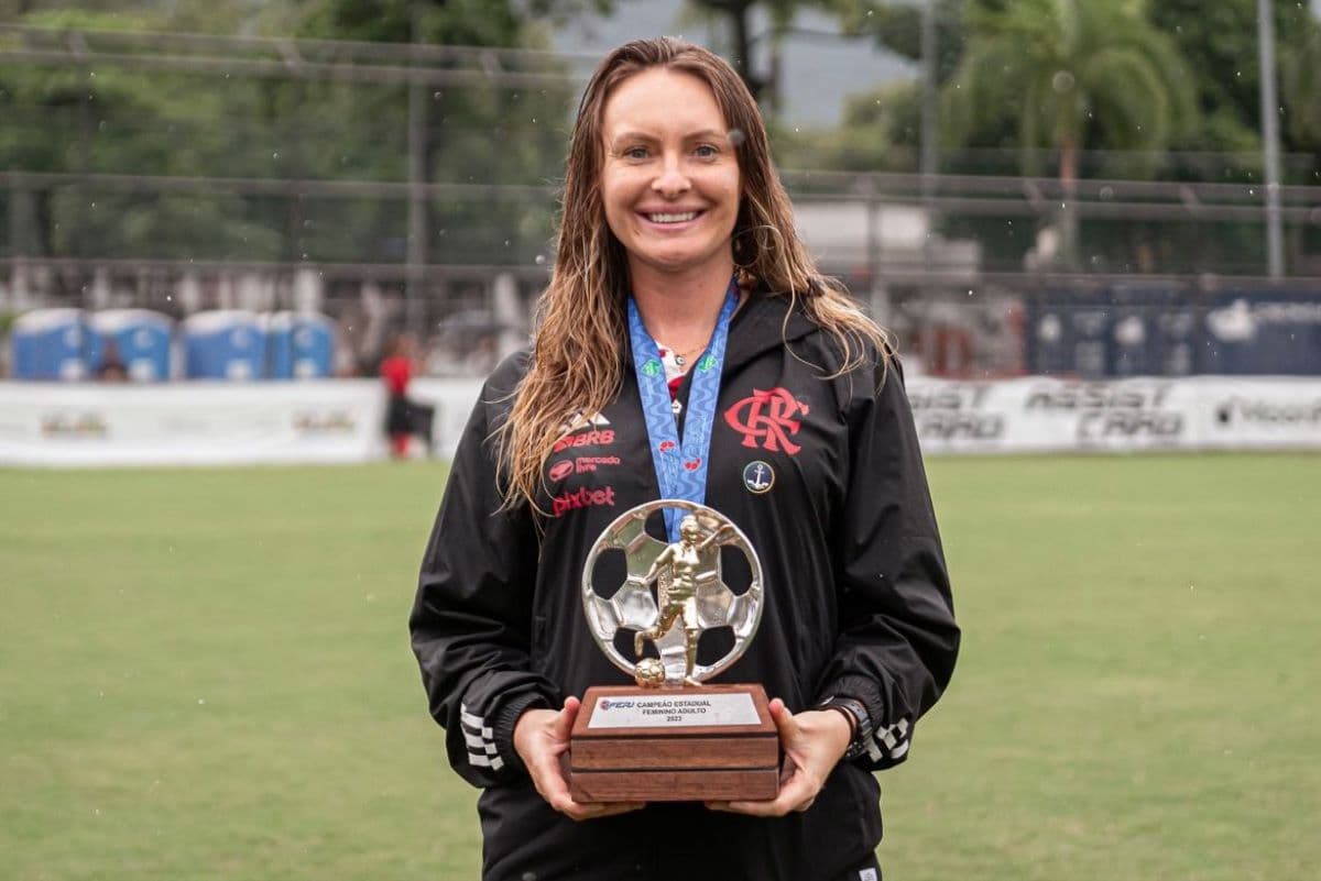 A auxiliar técnica está exibindo o troféu do campeonato carioca feminino, a ela sorri e está debaixo de chuva, a medalha está no pescoço e ao fundo está o campo da Gávea
