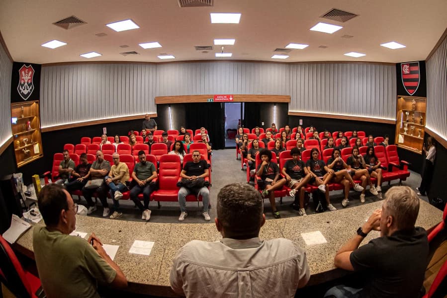 Na foto estão Maurício Salgado, André Rocha e Fábio Palmer sentados de frente para o auditório, nas cadeiras da sala de reuniões estão as jogadoras do time profissional e integrantes da comissão