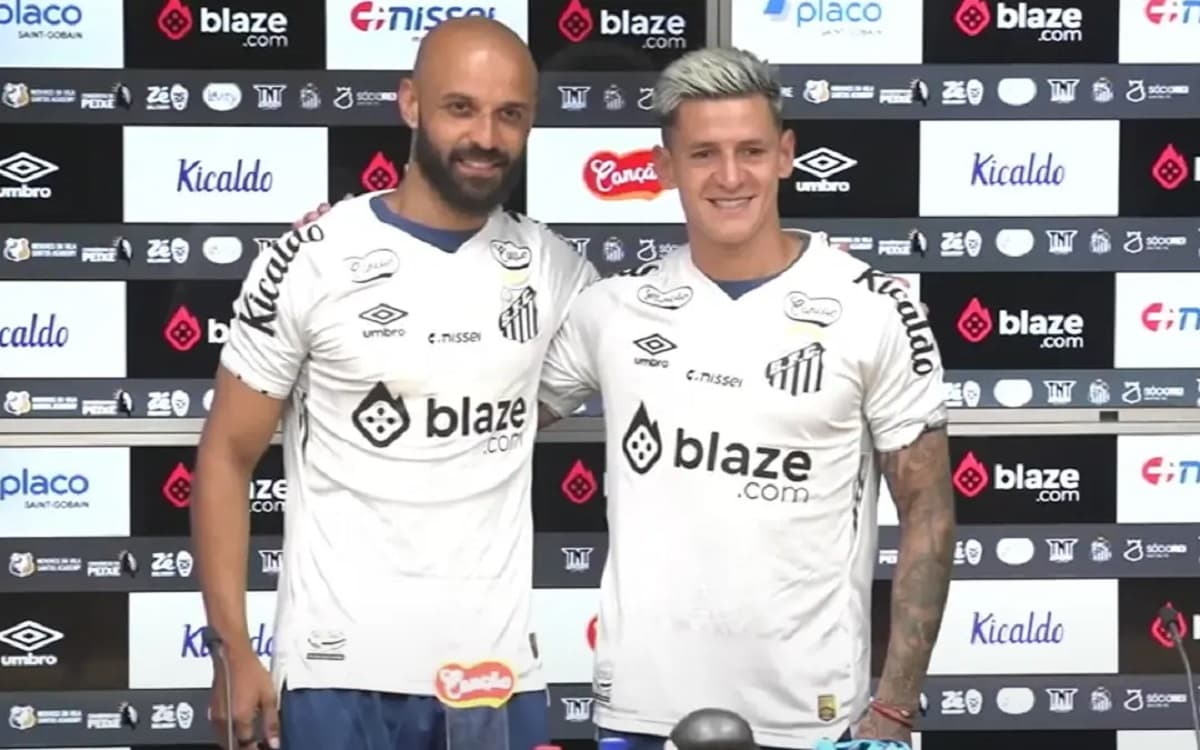 Vestindo a camisa branca do Santos, Thaciano e Léo Godoy posam para fotos com os patrocinadores do clube ao fundo