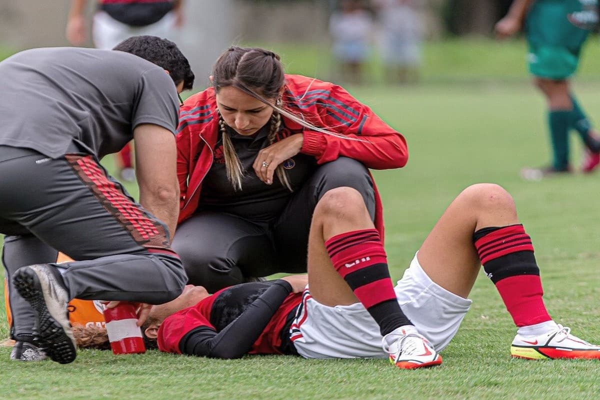 Thais está agachada realizando um atendimento a uma jogadora que caiu em campo