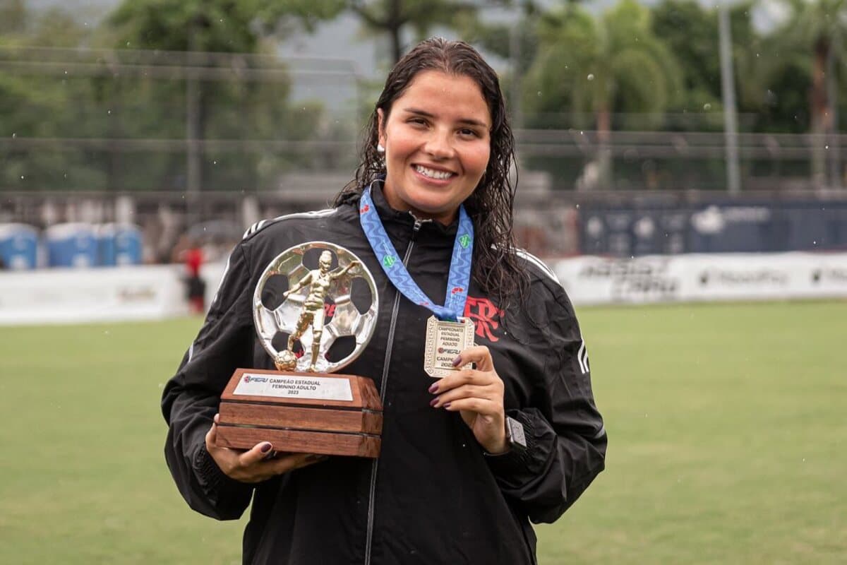 A Psicóloga do Flamengo feminino está na chuva, de casaco e sorrindo, ela segura a taça e medalha do campeonato carioca