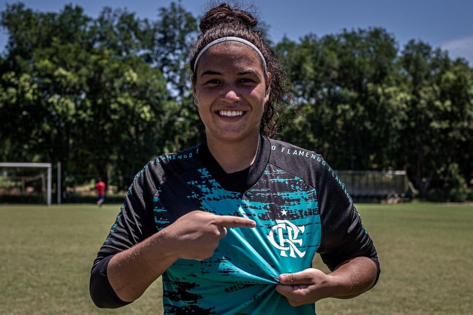 A goleira Yasmin está usando o uniforme azul, sorrindo e apontando para o escudo do Flamengo