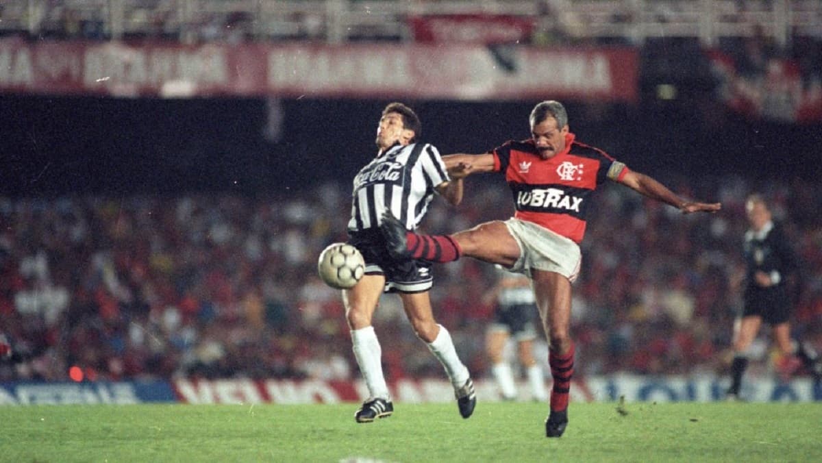 Com a camisa rubro-negra do Flamengo, Junior disputa bola no campo com jogador do Botafogo