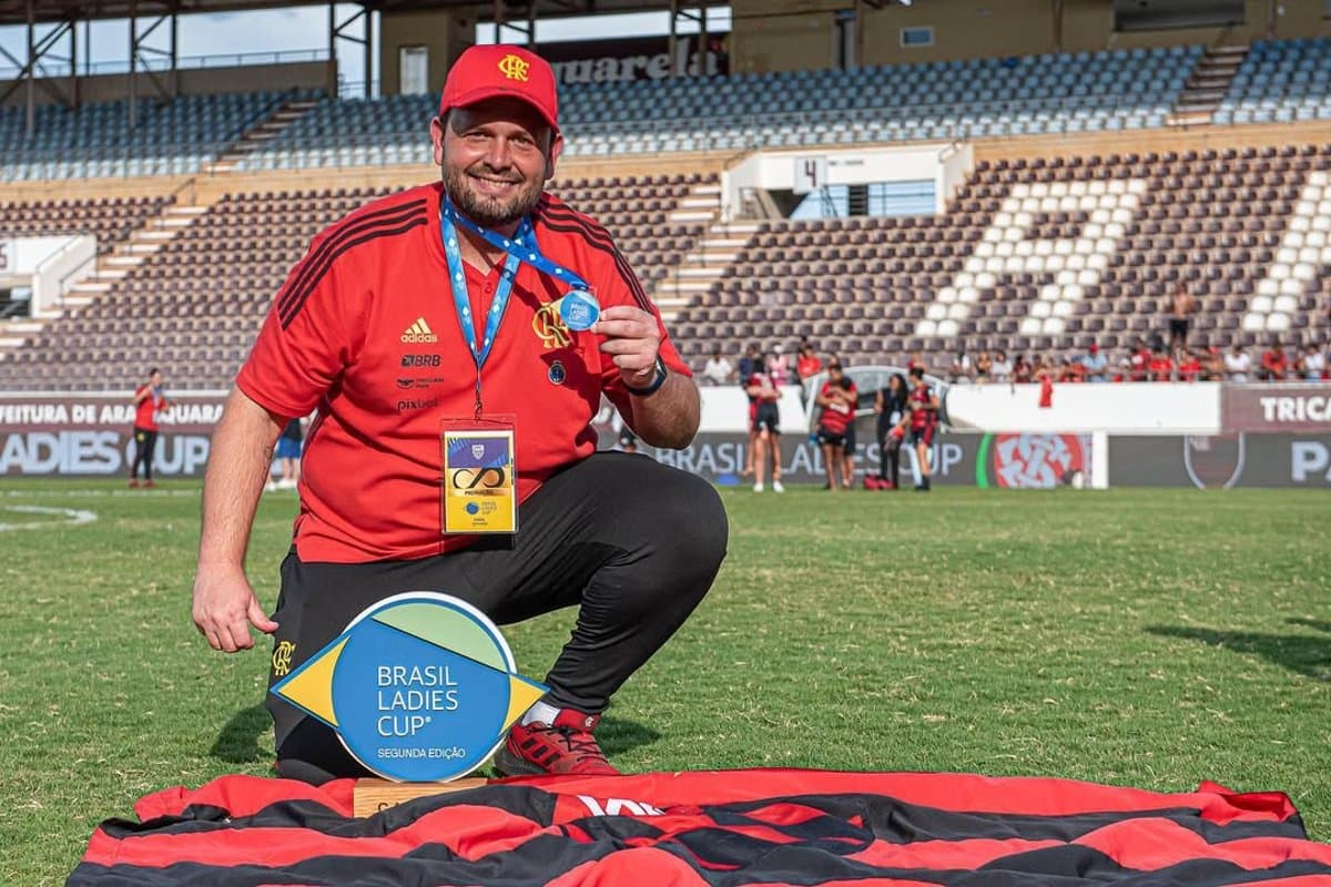 Felipe está com o uniforme da comissão técnica e boné, ele está agachado, segurando a medalha da Ladies cup e a sua frente tem uma bandeira do Flamengo esticada com o troféu de campeão em cima