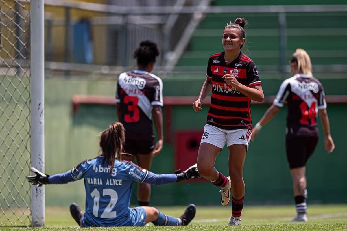 Laysa comemora gol, ela está sorrindo, na foto também aparecem três jogadoras do vasco: a goleira que está sentada no chão de costas e duas outras atletas andando na direção oposta de Laysa