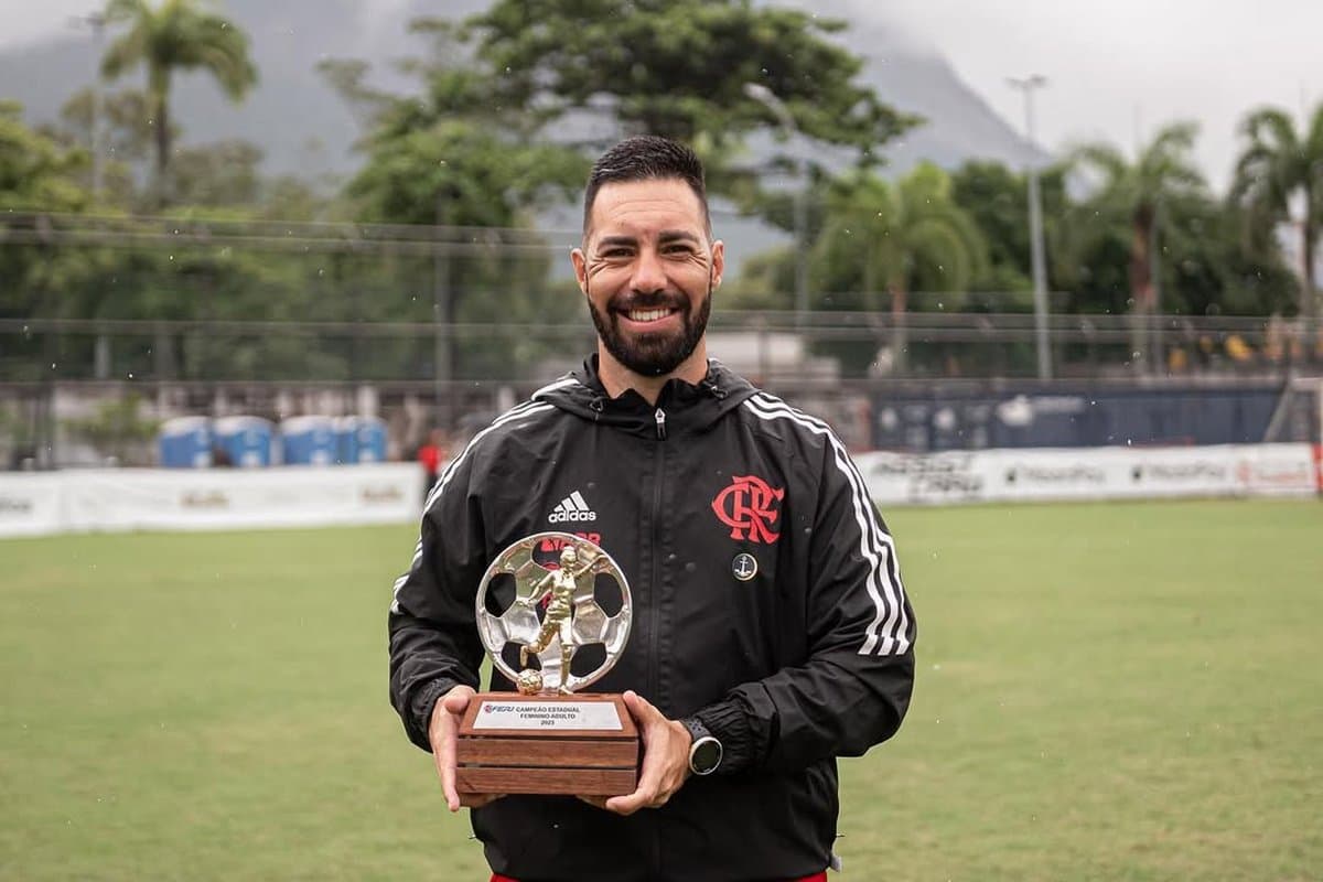 Felipe está sorrindo, vestindo um casaco do Flamengo, na chuva, segurando a taça do carioca feminino adulto