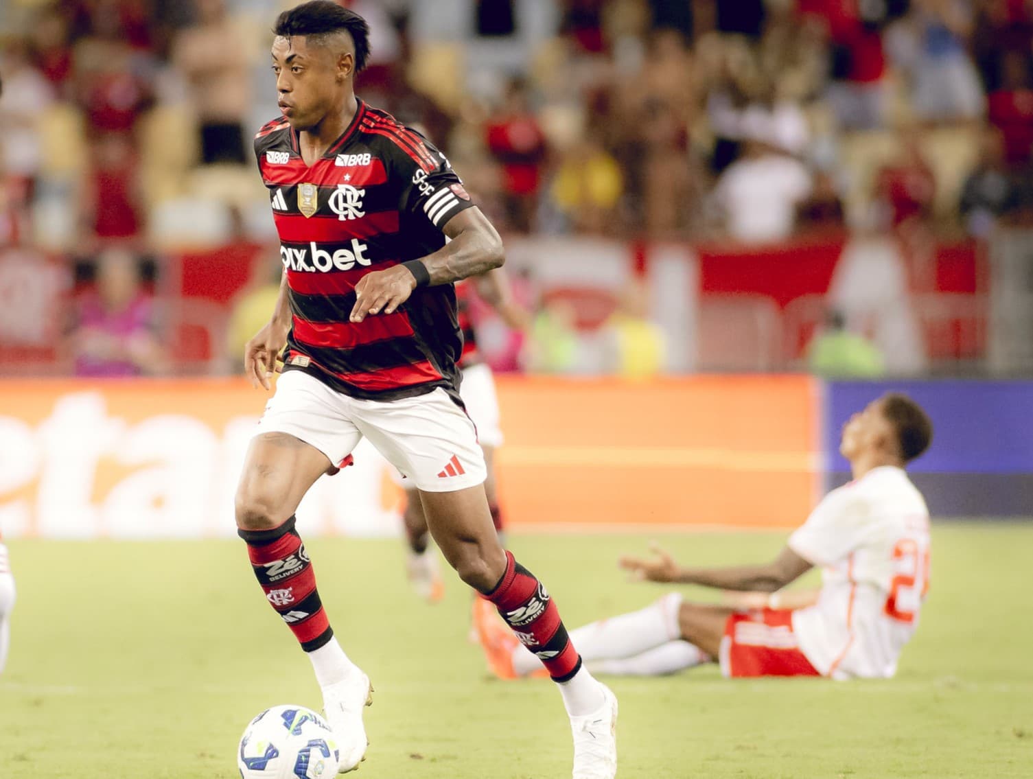 Bruno Henrique carrega a bola para o ataque; ao fundo, jogador do Internacional lamenta sentado no chão