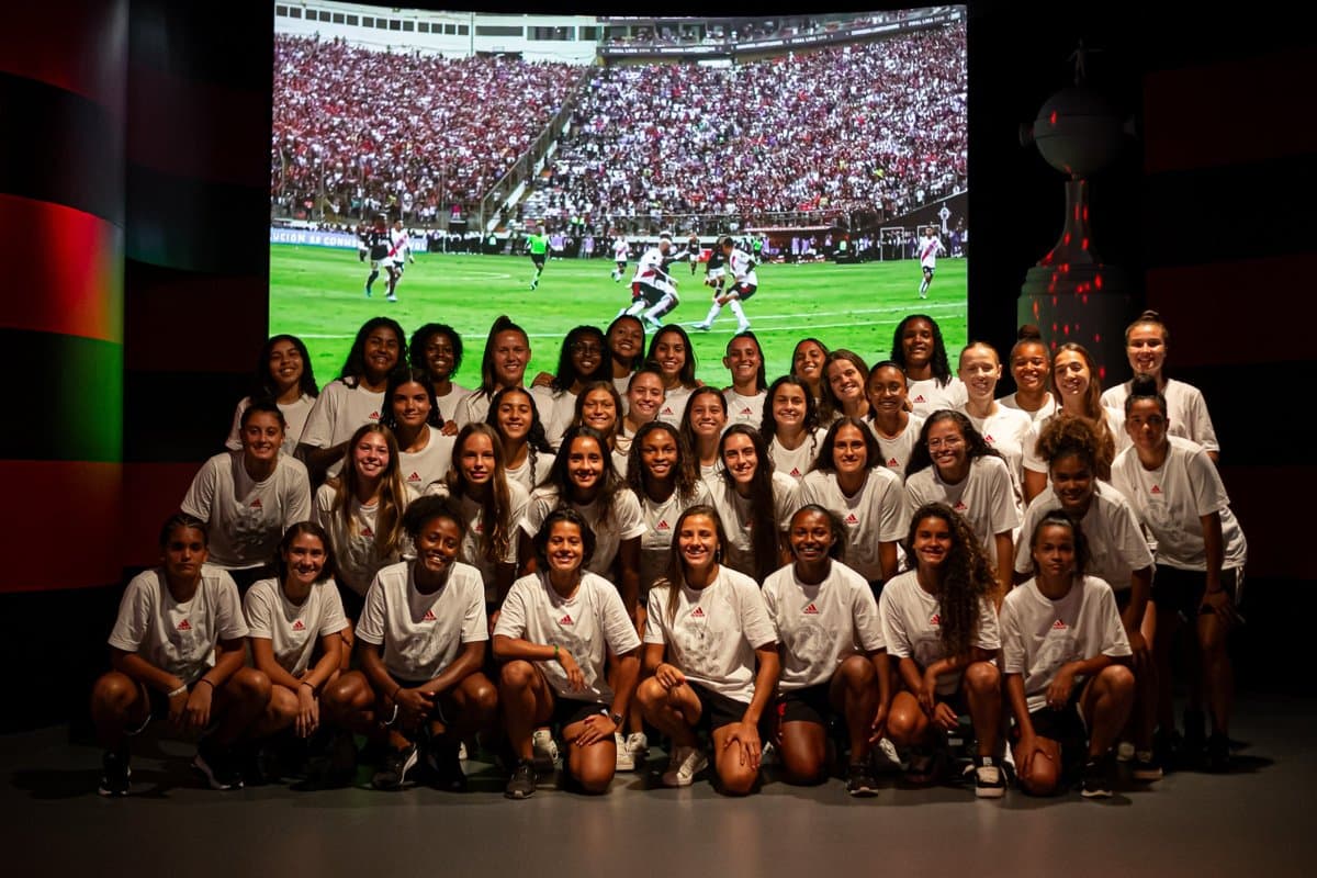 Todo o elenco do Sub-20 está posicionado a frente de um telão no museu da Gávea, as atletas estão sorrindo e com uma blusa branca que possui a logo da adidas em vermelho e abaixo o CRF em prata