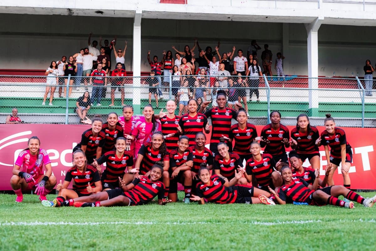 O time está reunido no gramado, posando para a foto e ao fundo torcedores rubro-negros celebram a vitória em Laranjeiras