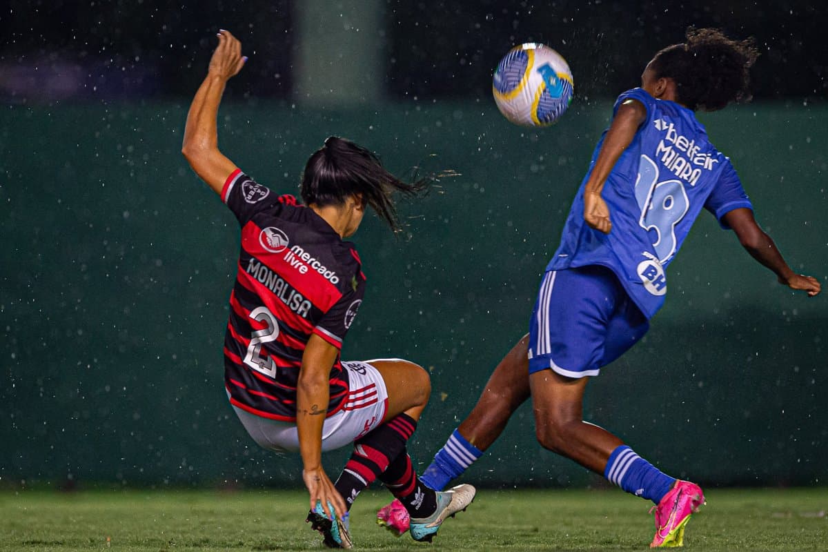 Monalisa e Maiara disputam a bola em Flamengo e Cruzeiro no Luso Brasileiro, Monalisa está mais próxima ao chão, como se estivesse dando um carrinho, já a jogadora do Cruzeiro está de pé tentando dominar a bola