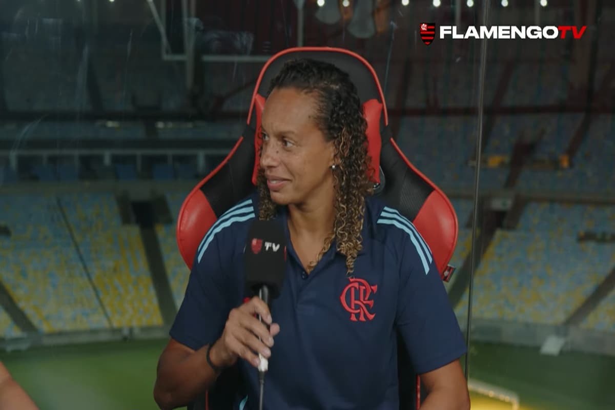 Rosana está no estúdio da Flamengo TV, segurando o microfone, veste o uniforme tradicional da comissão técnica do Rubro-Negro e ao fundo é possível ver campo e arquibancadas do Maracanã