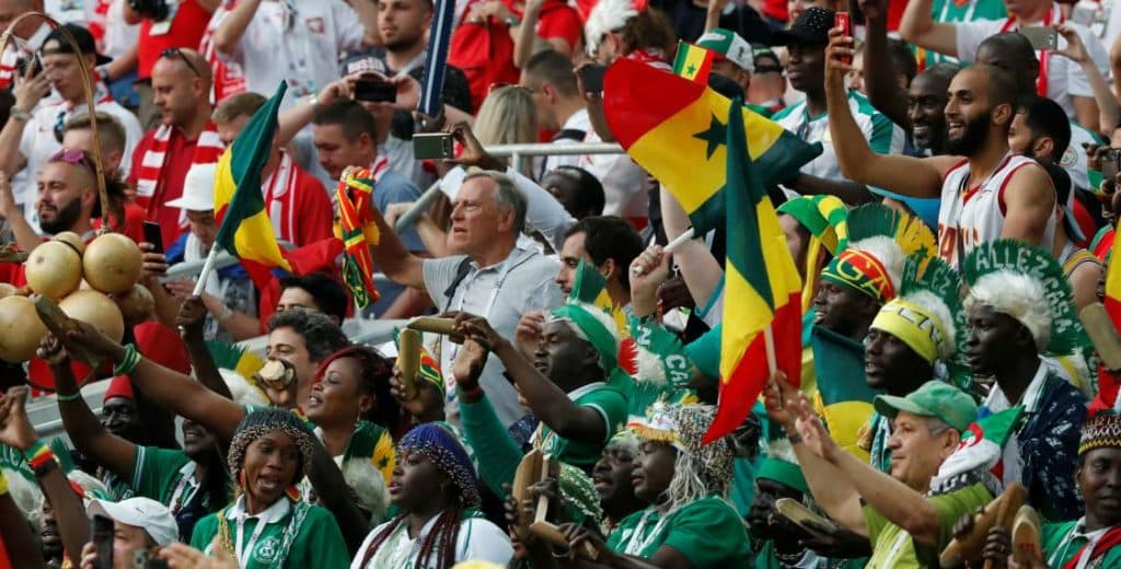 Torcedores africanos com bandeiras e camisas coloridas vibrando nas arquibancadas durante as Eliminatórias da Copa do Mundo 2026.