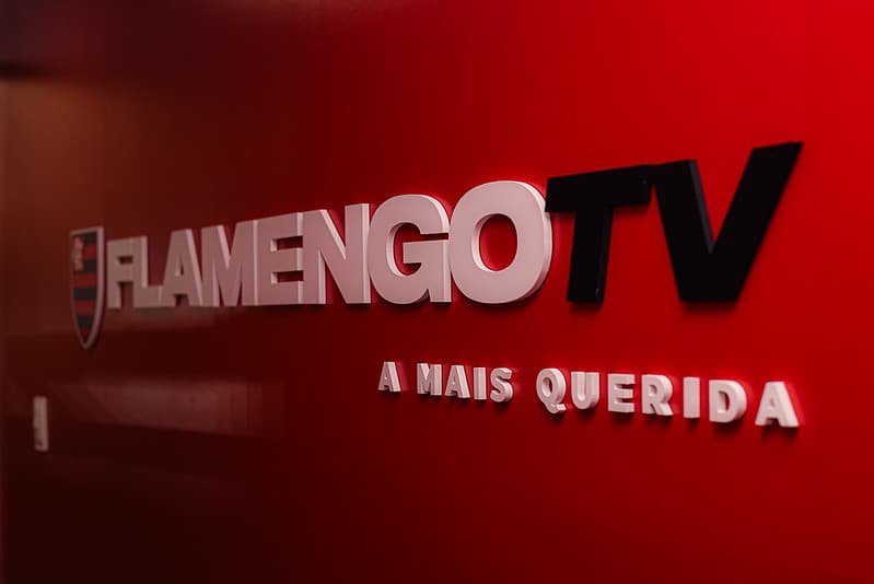 Logo da Flamengo TV com o escudo do Flamengo e o slogan “A Mais Querida” em parede vermelha.