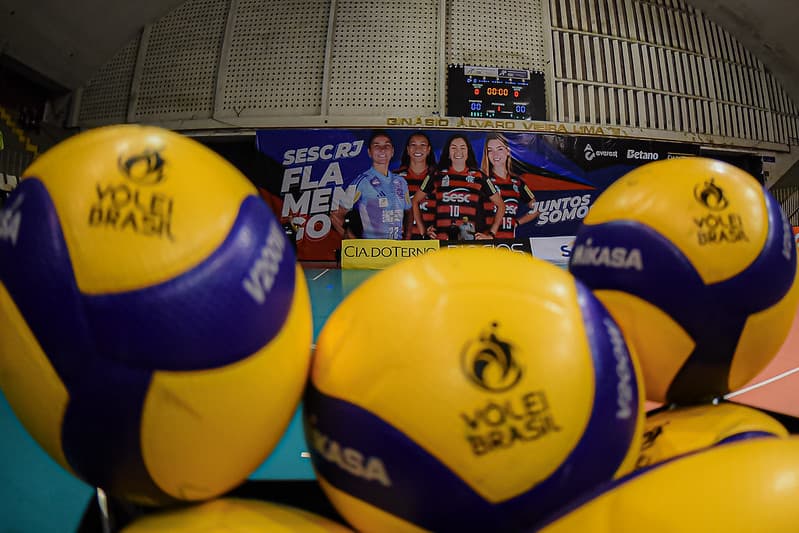 Bolas em primeiro plano, desfocadas, e o bunner do sesc flamengo com atletas no ginásio do tijuca ao fundo, focado.