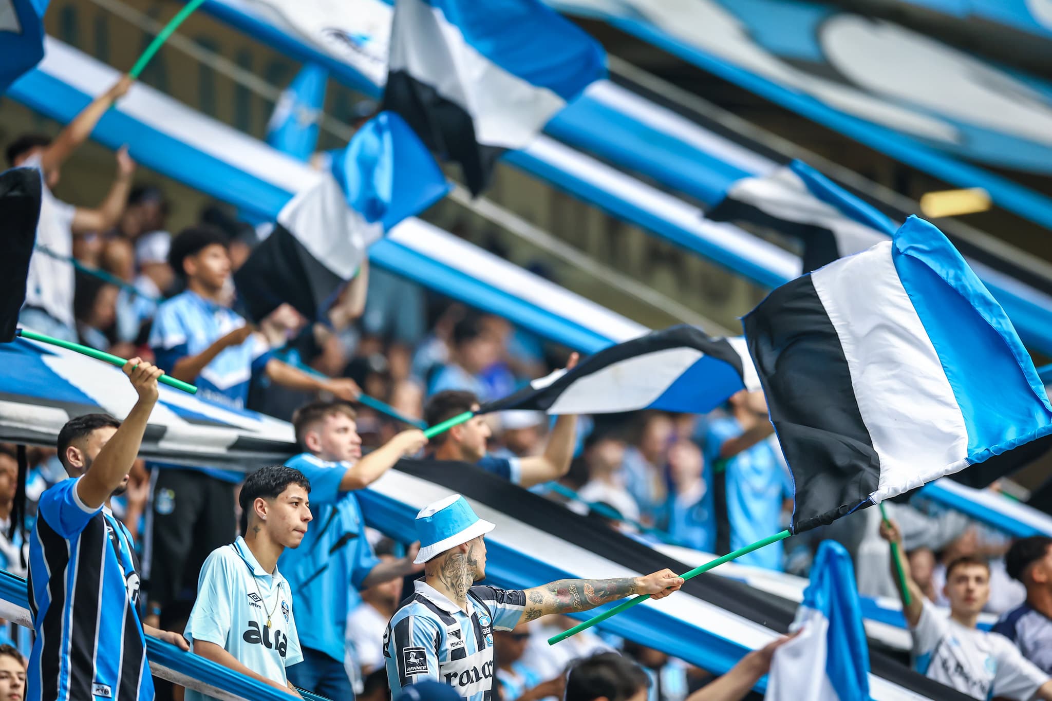 Torcida do Grêmio em jogo do clube na Arena, alguns torcedores seguram bandeiras com as cores do time: azul, preto e branco