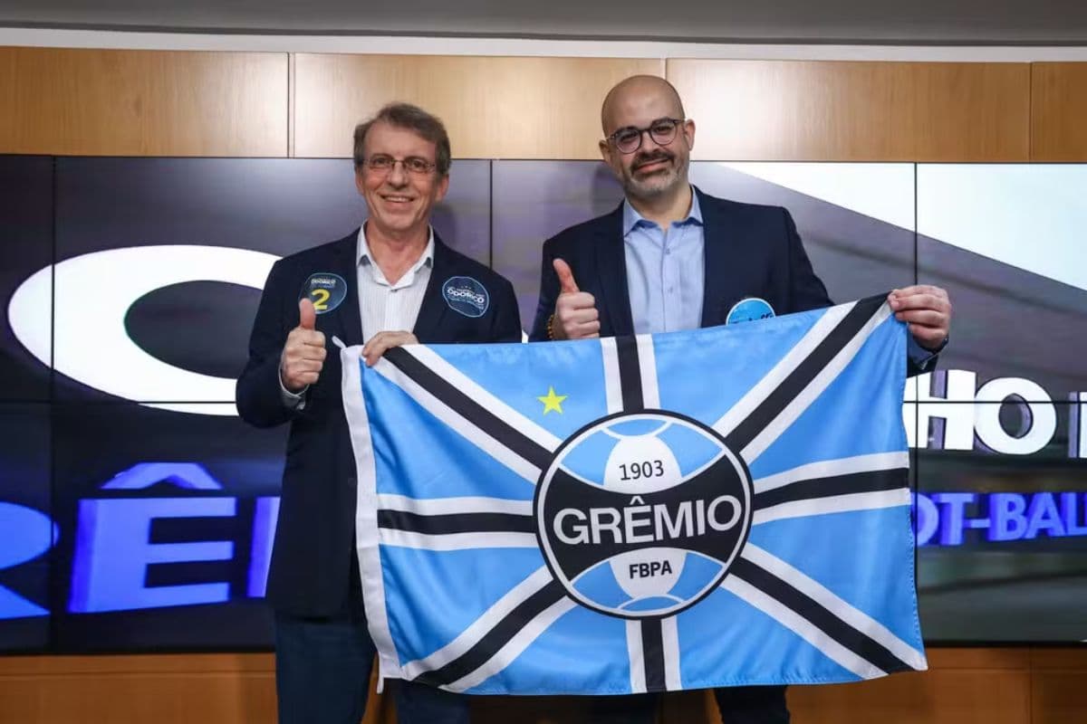 Odorico Roman e Paulo Caleffi, candidatos à presidência do Grêmio, posam juntos para foto segurando uma bandeira com o escudo e as cores do time