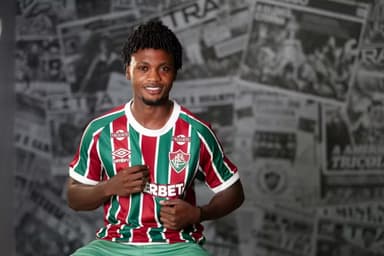 Jogador colombiano Santi Moreno posa para foto em sua apresentação no Fluminense. Ele sorri enquanto, com a mãos, dá foco no escudo do time tricolor