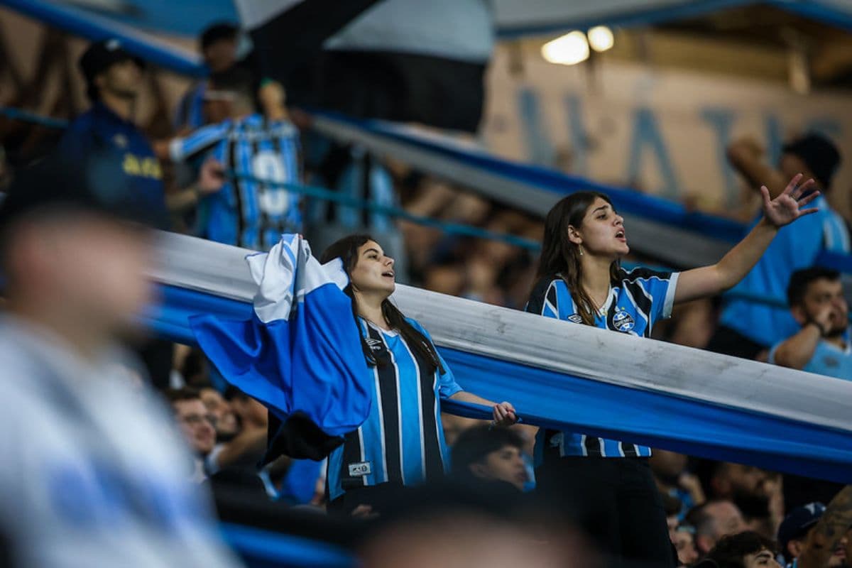 Imagem mostra torcedoras do Grêmio torcendo pelo clube em jogo do Campeonato Brasileiro na Arena do Grêmio. Uma delas segura uma bandeira do clube e a outra está com o braço levantado enquanto expressa apoio ao time