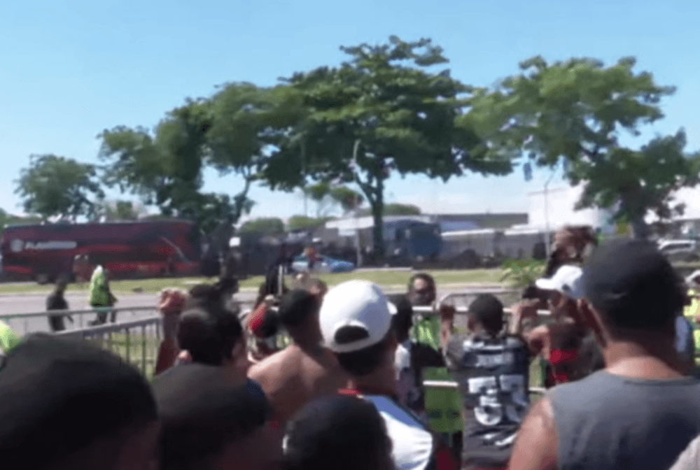 Torcedores do Flamengo empurram a grade de proteção, enquanto ônibus entra no Galeão ao fundo, longe da torcida no AeroFla