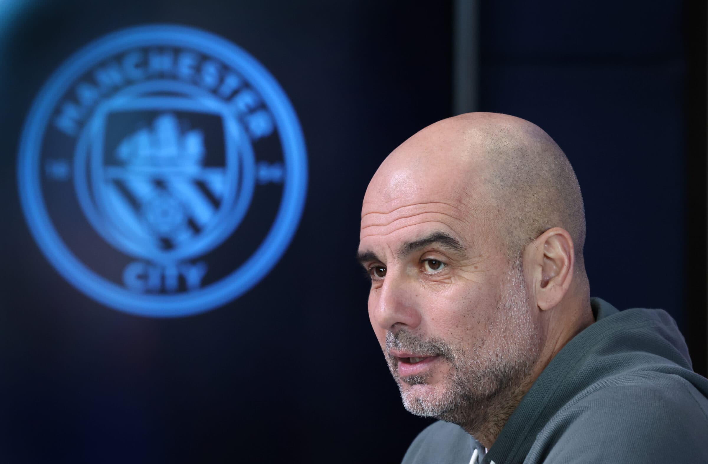 Técnico Pep Guardiola com escudo do Manchester City ao fundo
