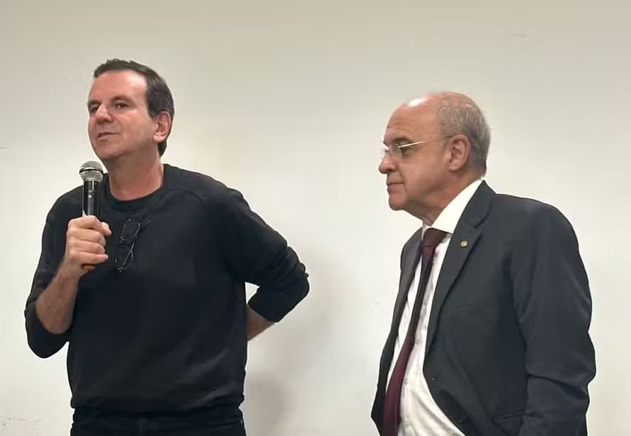 Eduardo Paes (à esquerda) discursa com microfone na mão e mão nas costas, enquanto Eduardo Bandeira de Mello (à direita) observa com semblante fechado