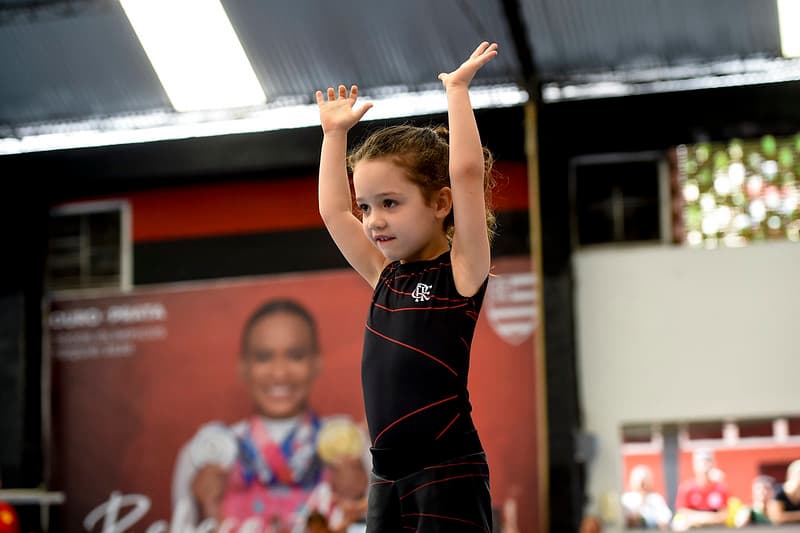 Ginasta infantil prepara pirueta; ao fundo painel de Rebeca Andrade