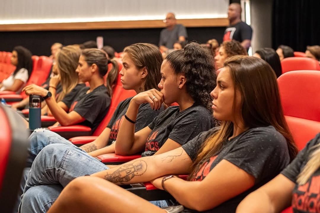a câmera está focada nas jogadoras, que estão sentados, olhando em direção ao palco, onde estão palestrando: Maurício Salgado, Fábio Palmer e André Rocha