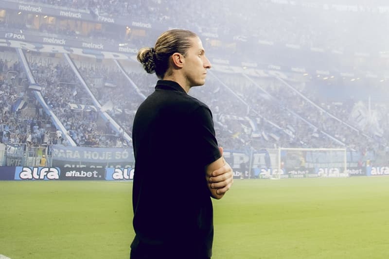 Filipe Luís de braços cruzados e semblante sério enquanto ao fundo torcida do Grêmio festeja entrada do time em campo na Arena do Grêmio