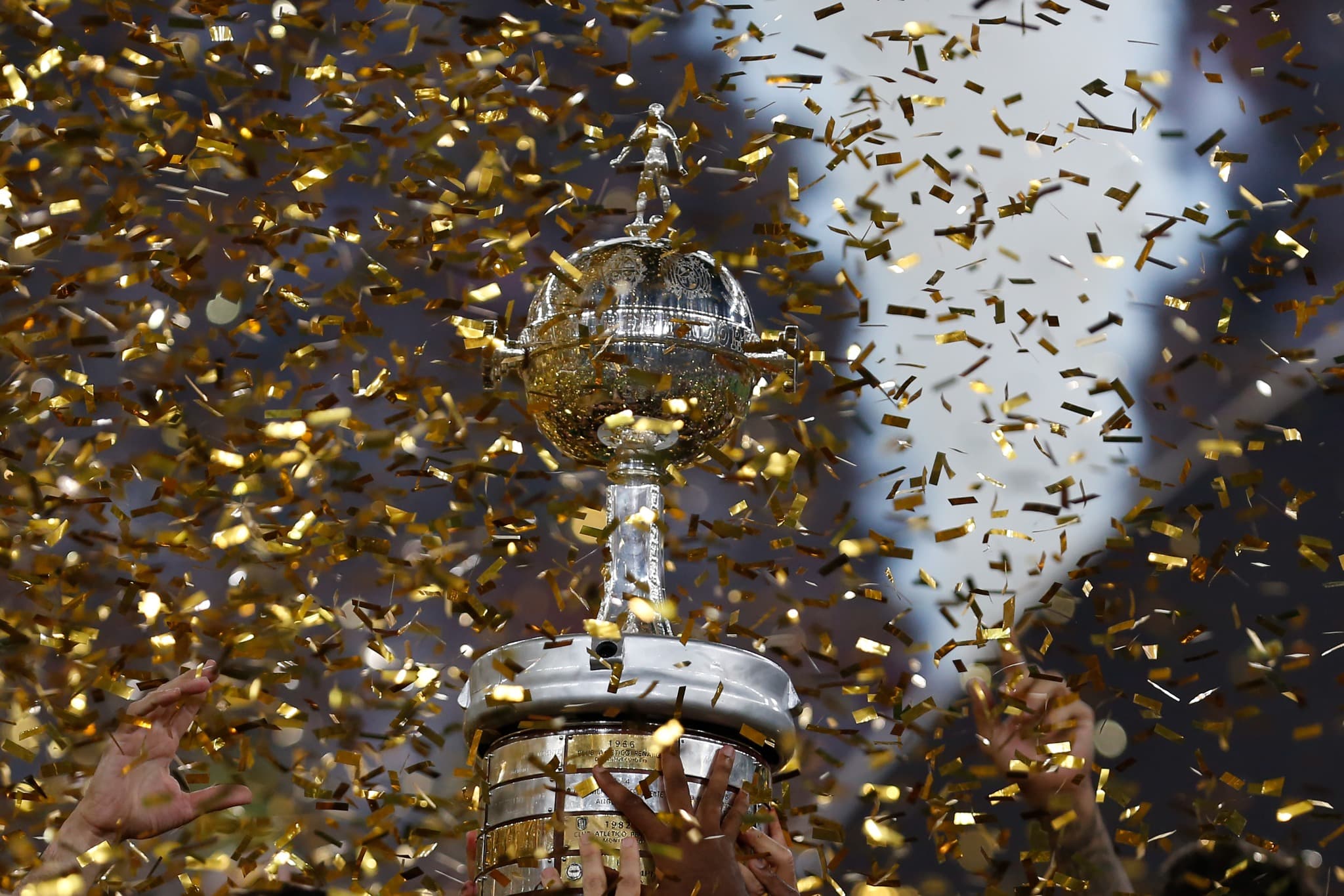 Taça da Copa Libertadores erguida em meio a papel picado dourado, simbolizando a grande final entre Flamengo e Palmeiras em 2025