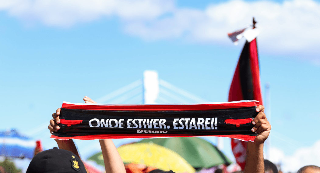 Torcedor do Flamengo ergue uma faixa escrita “Onde estiver, estarei!” durante o AeroFla, com bandeiras rubro-negras e céu azul ao fundo.