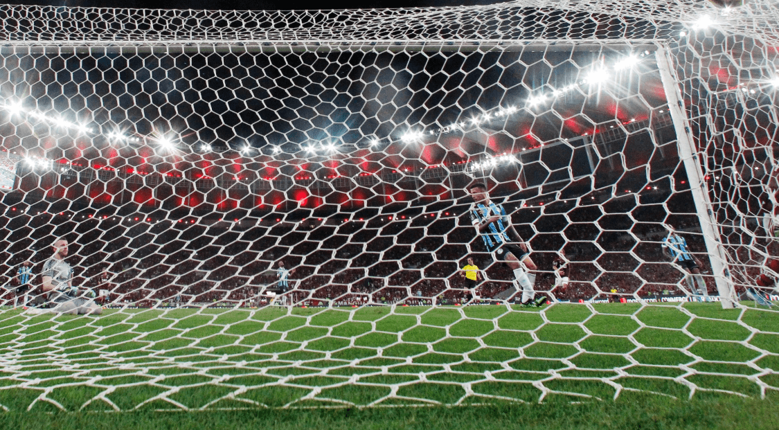 Rede vista de dentro do gol em jogo do Grêmio, com o goleiro e zagueiros ao fundo, simbolizando a força defensiva da equipe em temporadas recentes.