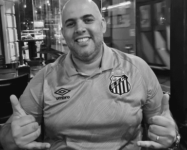 Marcão do Varanda usando camisa do Santos sentado em um restaurante, sorrindo e fazendo gesto com as mãos, em foto utilizada como homenagem.