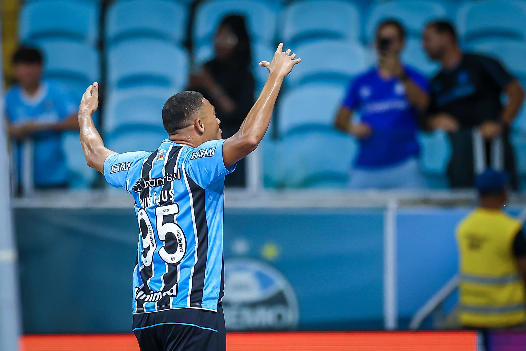 Carlos Vinicius com braços levantados e comemorando gol pelo Grêmio contra o Bragantino junto com a torcida na Arena